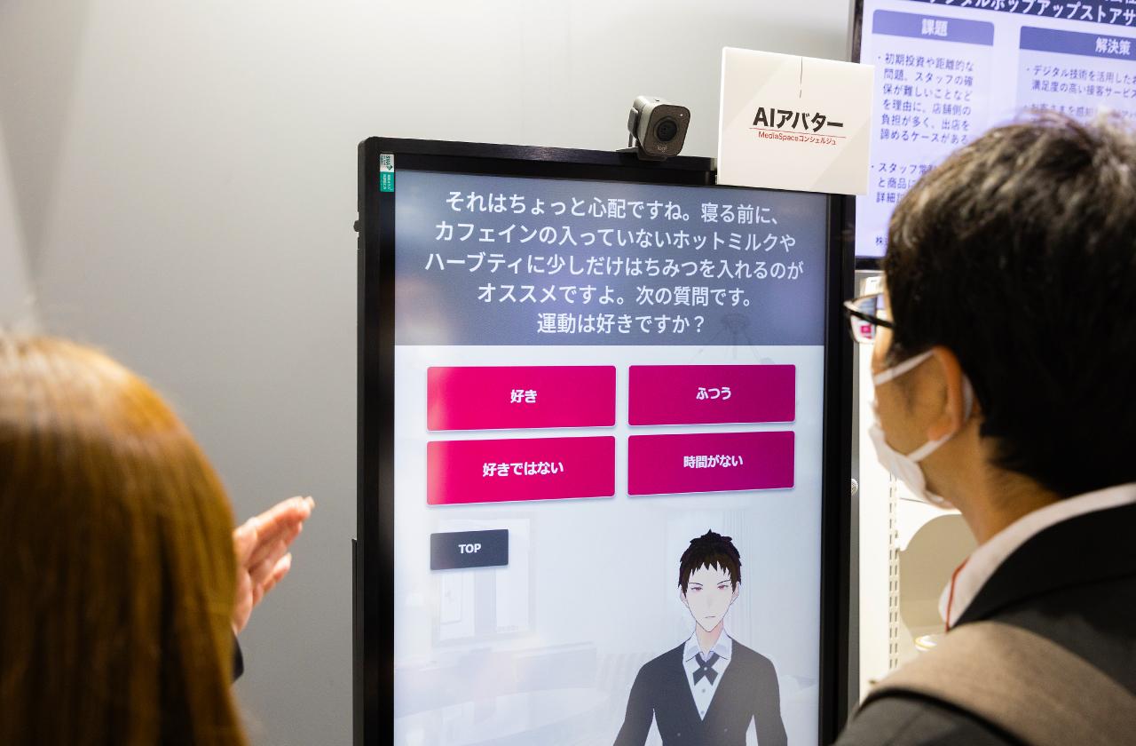画像: タッチパネルでAIアバターからの質問に答えると、音声とテキストで返答し、次の質問をしてくる。例えば「朝までぐっすり眠れますか？」という質問に対し「ぐっすり眠れない」を選択すると、「それはちょっと心配ですね。寝る前に、カフェインの入っていないホットミルクやハーブティに少しだけはちみつを入れるのがオススメですよ」というように、回答に応じたコメントでワンクッション入れてから次の質問に進む。コメントは、サイネージ上部に設置されたAIカメラで回答者の表情や性別を認識し、AIが選択している。購買客との会話のシナリオは約50パターンあり、より細かく分岐させることでさらに増やすこともできる