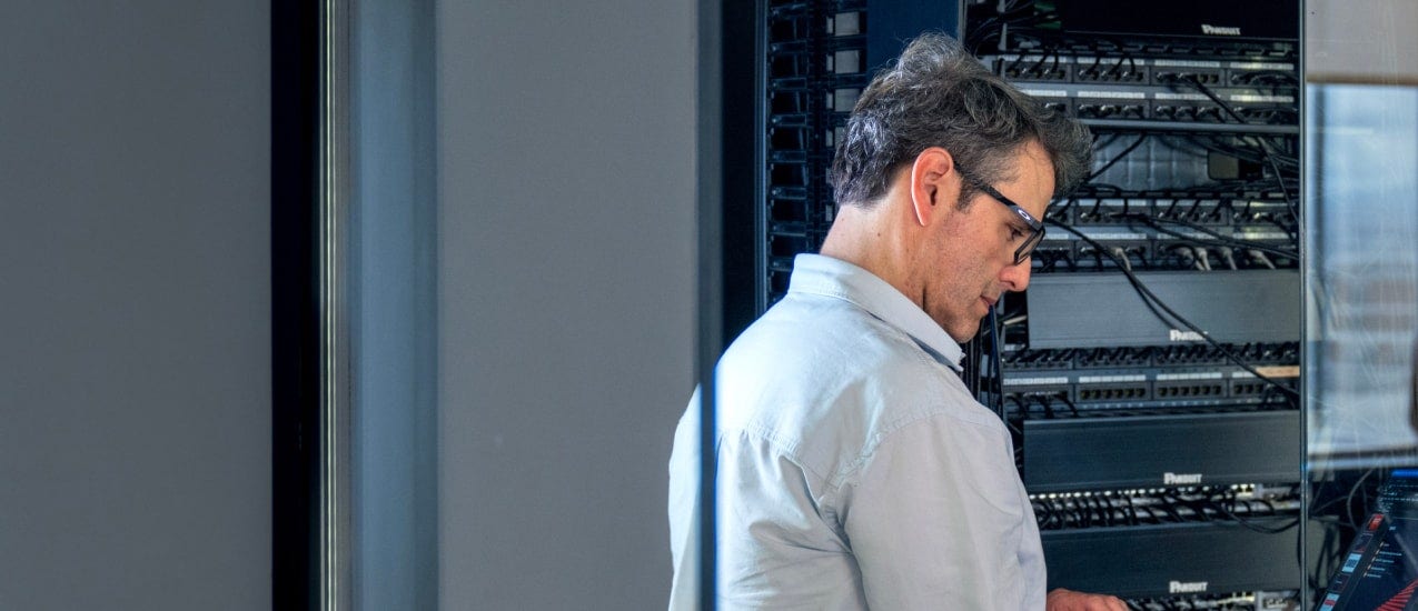 A person wearing glasses and light-coloured shirt in front of a server rack